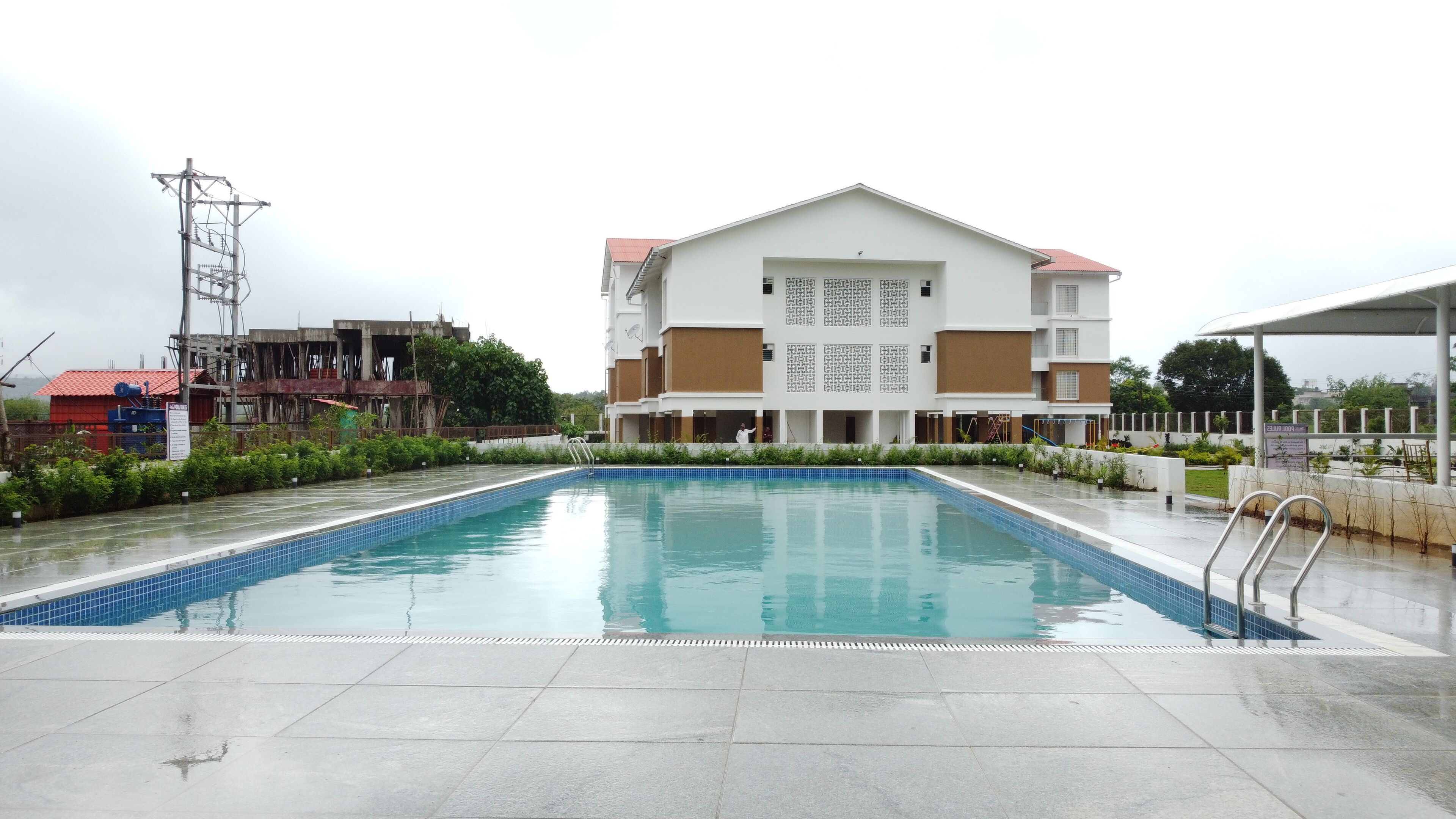 A swimming pool with a tiled deck, a multi-story building with a red roof in the background, and a utility tower to the left.