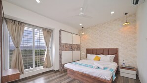 A modern hotel room with a large bed, a nightstand, a ceiling fan, and a window with curtains. -  Ovi Resort and Spa (Mawal)