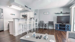 Una sala de estar moderna con área de cocina, muebles blancos, televisor y una mesa de centro con controles remotos.
