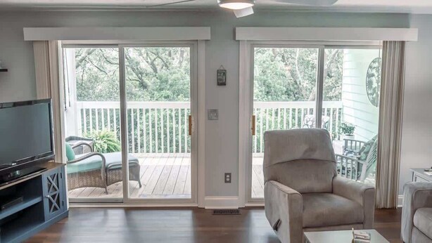 Una sala de estar con televisión, ventilador de techo, puerta corrediza de vidrio a una terraza y un sillón color beige.