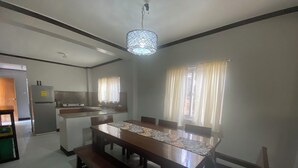 A dining area with a long wooden table, chairs, and a patterned table runner. There is a kitchen area with a refrigerator and a sink in the background.