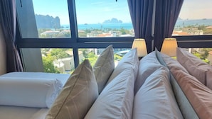 A hotel room with a large window offering a view of the sea and distant islands. There are multiple pillows and a bed with a white bedspread. - B303- 1 Bedroom Condo With Sea View, Ao Nang Beach (Krabi)