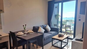 A modern living room with a dining table set for two, a sofa with cushions, and a view of the city through large windows. - B303- 1 Bedroom Condo With Sea View, Ao Nang Beach (Krabi)