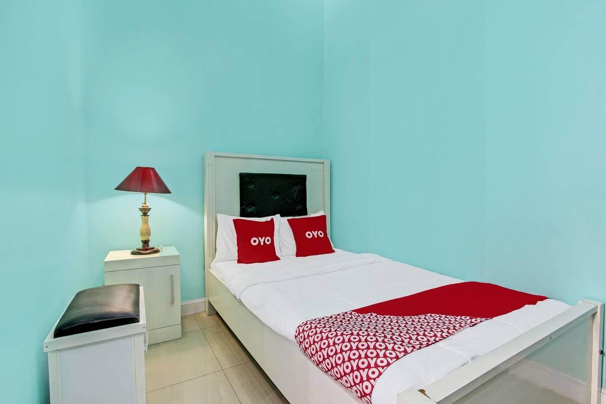 A single-bed room with a white bed, red pillows, a red throw blanket, a bedside table with a lamp, and a small white cabinet.