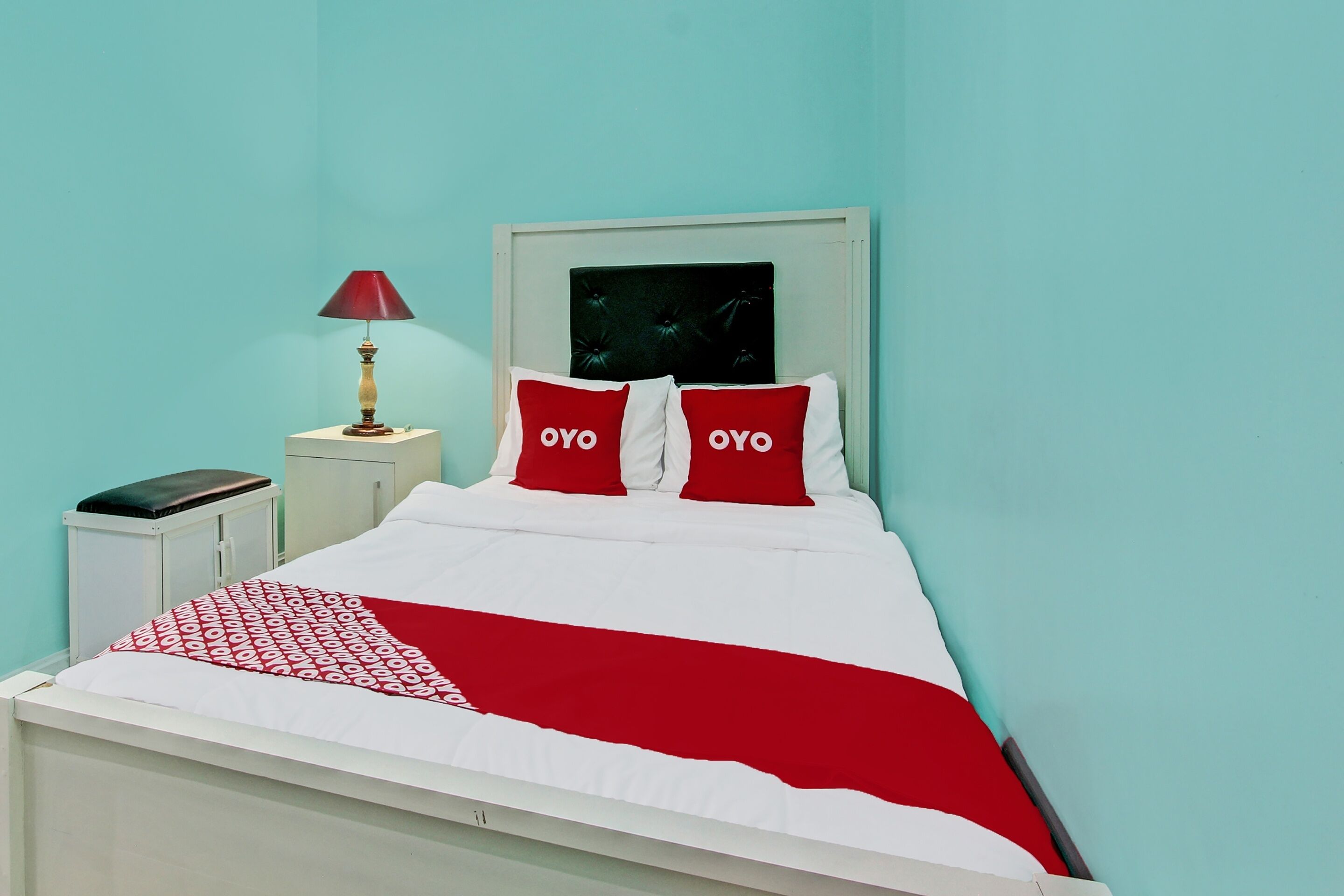 Un lit avec une literie blanche et rouge, deux oreillers rouges ornés du mot « OYO », une lampe rouge posée sur une table de chevet et une armoire blanche.