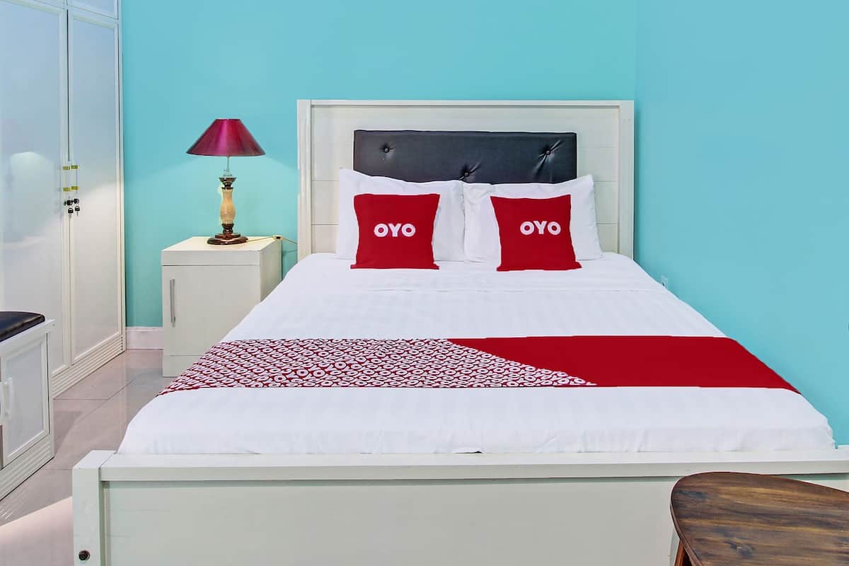 A neatly made bed with white bedding and two red pillows with the word "OYO" on them, a small bedside table with a lamp, and a wardrobe.