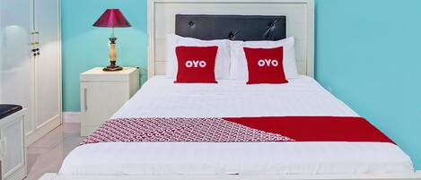 Un lit bien fait, avec une literie blanche et deux oreillers rouges ornés du mot « OYO », une petite table de chevet avec une lampe et une armoire.