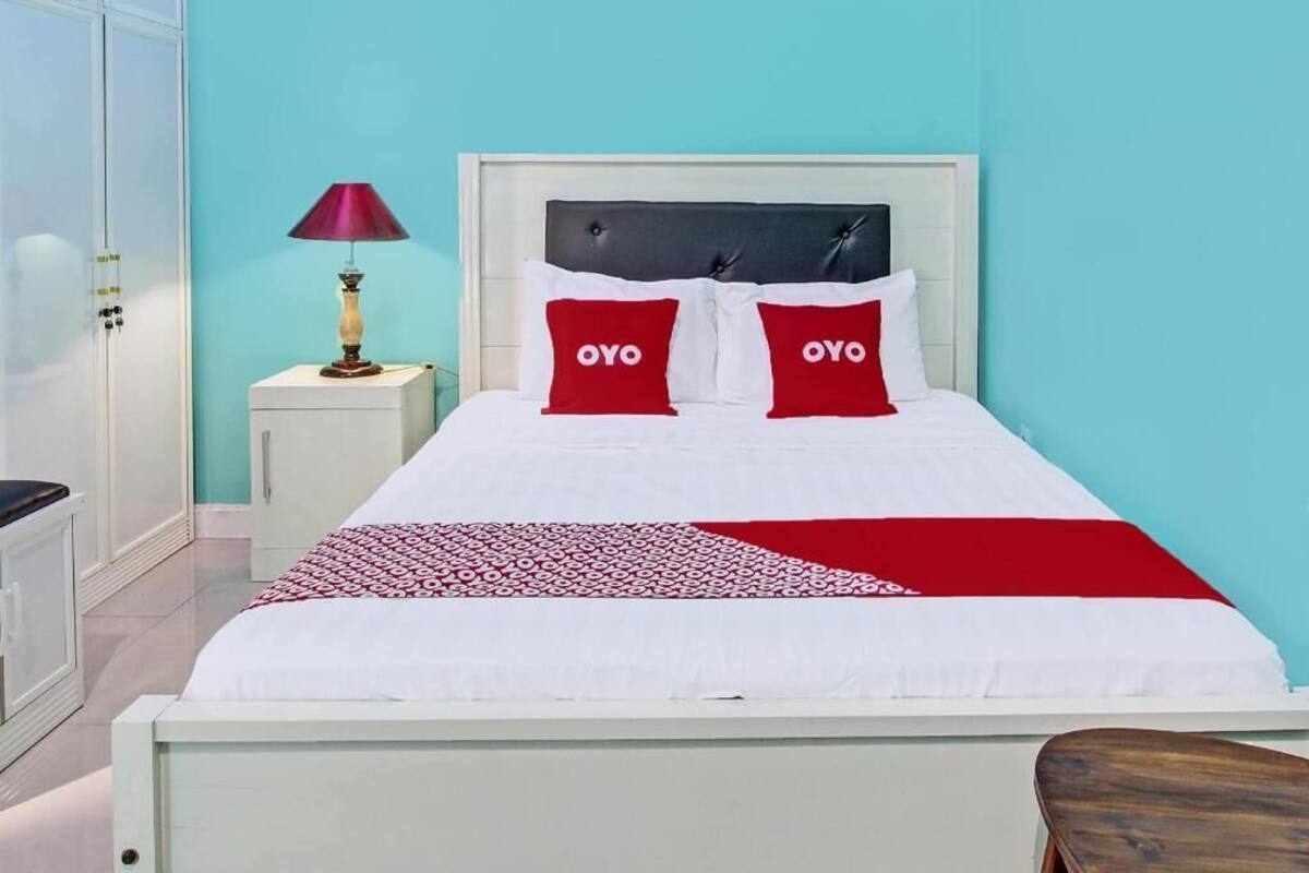 A bed with white bedding, two red pillows with 'OYO' text, a red and white patterned throw, a white nightstand with a lamp, and a wooden bench.
