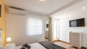 Une chambre à coucher comprenant un lit, une lampe de chevet, une commode, un téléviseur fixé au mur et une fenêtre avec des stores.
