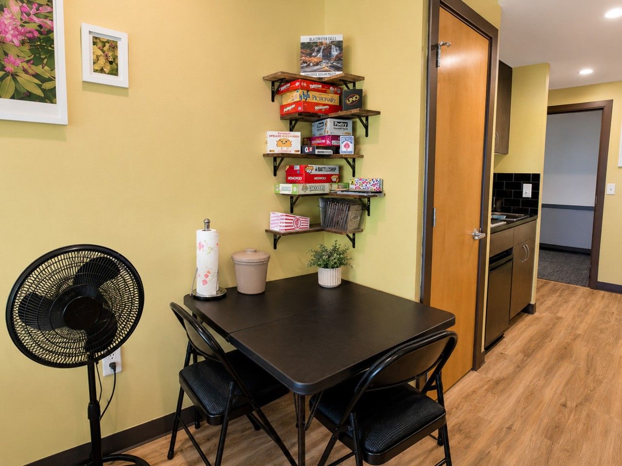 Un petit coin repas comprenant une table, des chaises, un ventilateur et une étagère avec des jeux de société.