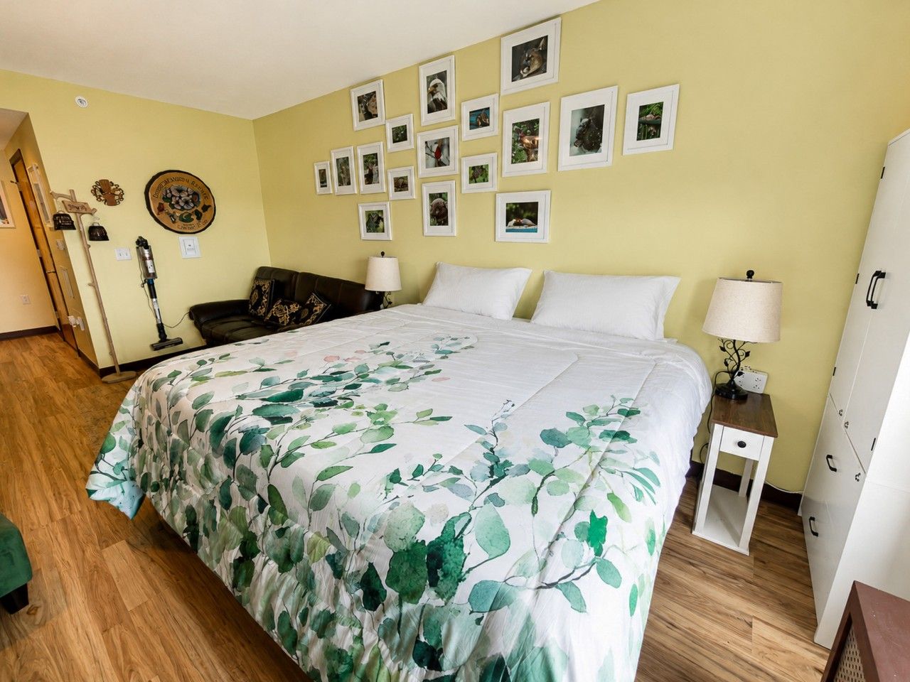 Une chambre à coucher avec un lit, une table de chevet, une lampe, un mur orné de tableaux encadrés et une porte.