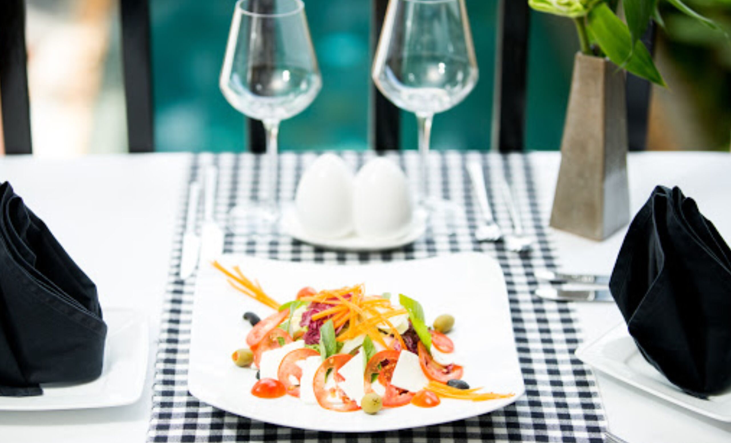 A dining table with a checkered tablecloth, a white plate with a salad, two glasses of water, and a vase with a plant.