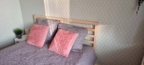 Ein Bett mit floralem Überwurf, zwei rosafarbene Kissen, ein kleiner Topfpflanze und ein Holz-Kopfteil.