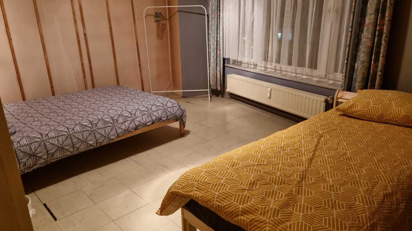 Ein Zimmer mit zwei Betten, einem Heizkörper, einem Fenster mit Vorhängen und einem Holzschrank.
