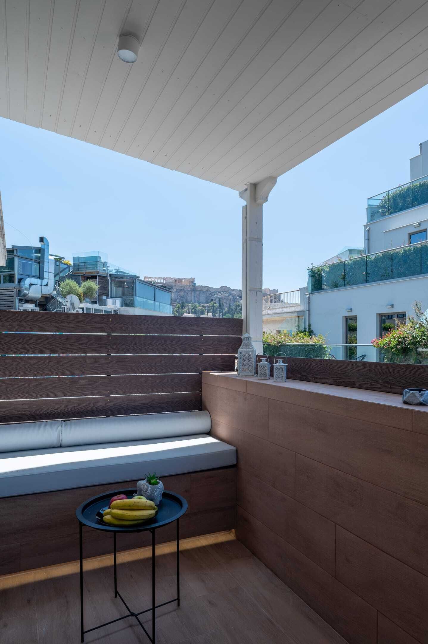 Un área de estar moderna al aire libre con un banco de madera y una mesita, con vistas a un paisaje urbano con edificios y al mar.