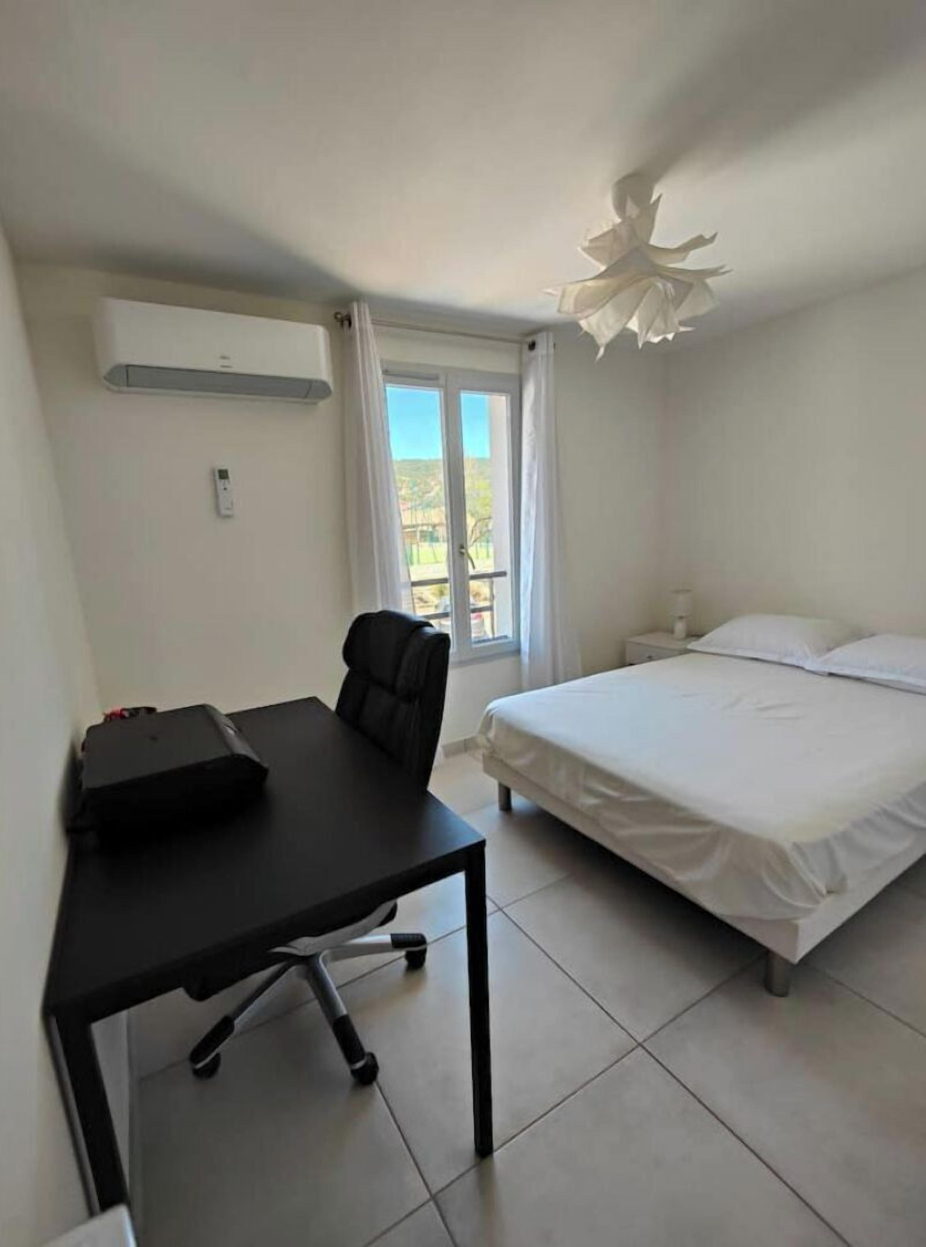 Een slaapkamer met een bed, een bureau, een stoel, een nachtkastje en een raam met gordijnen.