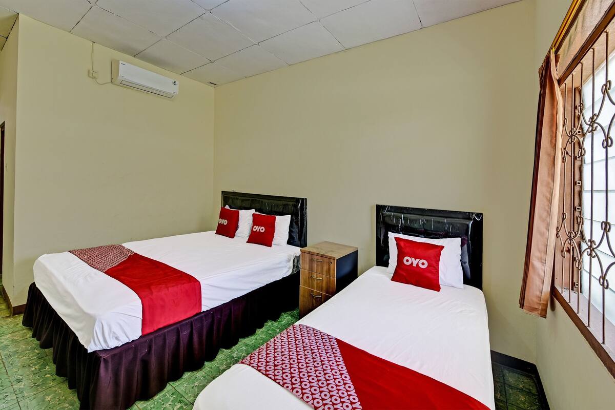 Ein Hotelzimmer mit zwei Betten, jeweils mit roter und weißer Bettwäsche und Kissen, einem hölzernen Nachttisch und einem Fenster mit Metallgitter.