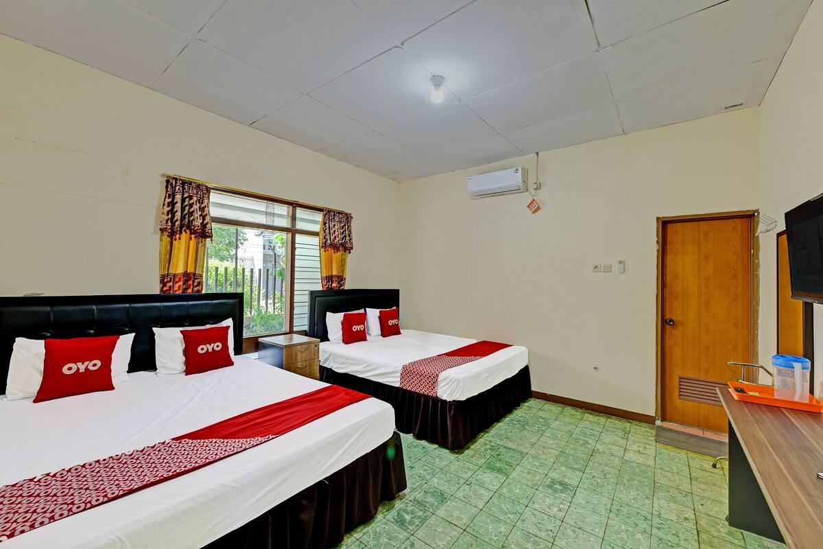 Ein Hotelzimmer mit zwei Betten, jeweils mit rot-weißer Bettwäsche, einem Holzkopfende und einem mit Vorhängen versehenen Fenster.