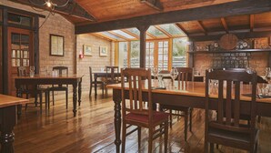 A rustic dining area with wooden tables and chairs, a brick wall, and a stone fireplace. - Danseys Pass Hotel (Kyeburn Diggings)