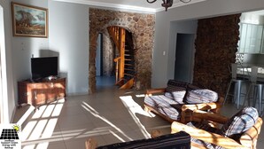 Un salon doté d’une cheminée en pierre, d’un escalier en bois, d’un téléviseur posé sur un meuble en bois, et d’un coin repas avec des chaises en métal.