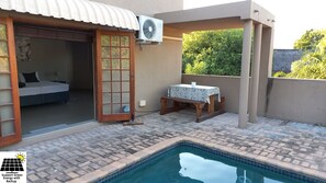 A pool area with a small table and bench, a fan, and a room with a bed and a sliding door. - SnL Guesthouse at Visarend (Nkomazi)