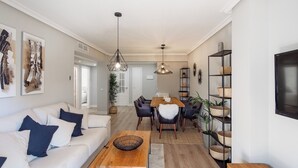 En moderne stue med sofa, sofabord, spisebord og stole. Et fjernsyn er monteret på væggen, og der står en plante i hjørnet.