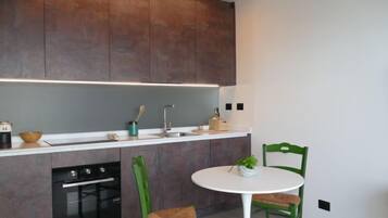 Una cocina moderna con armarios de madera oscura, encimera blanca, horno empotrado y una mesa redonda blanca con dos sillas verdes.