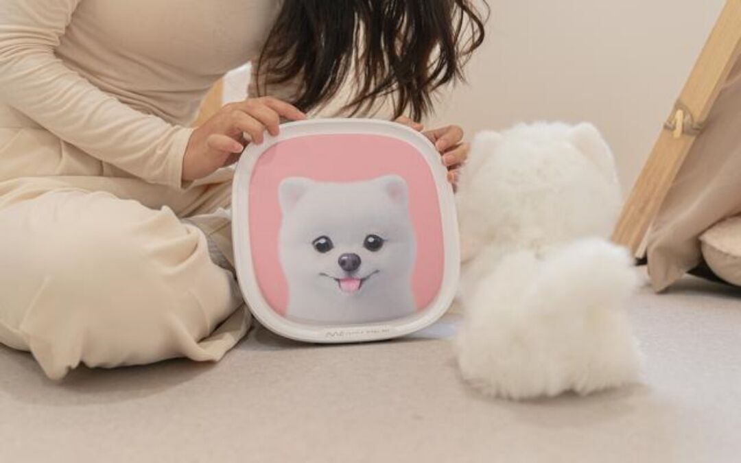 Una persona tiene in mano un quadro incorniciato con il ritratto di un cane, seduta a terra accanto a un cane bianco e soffice.