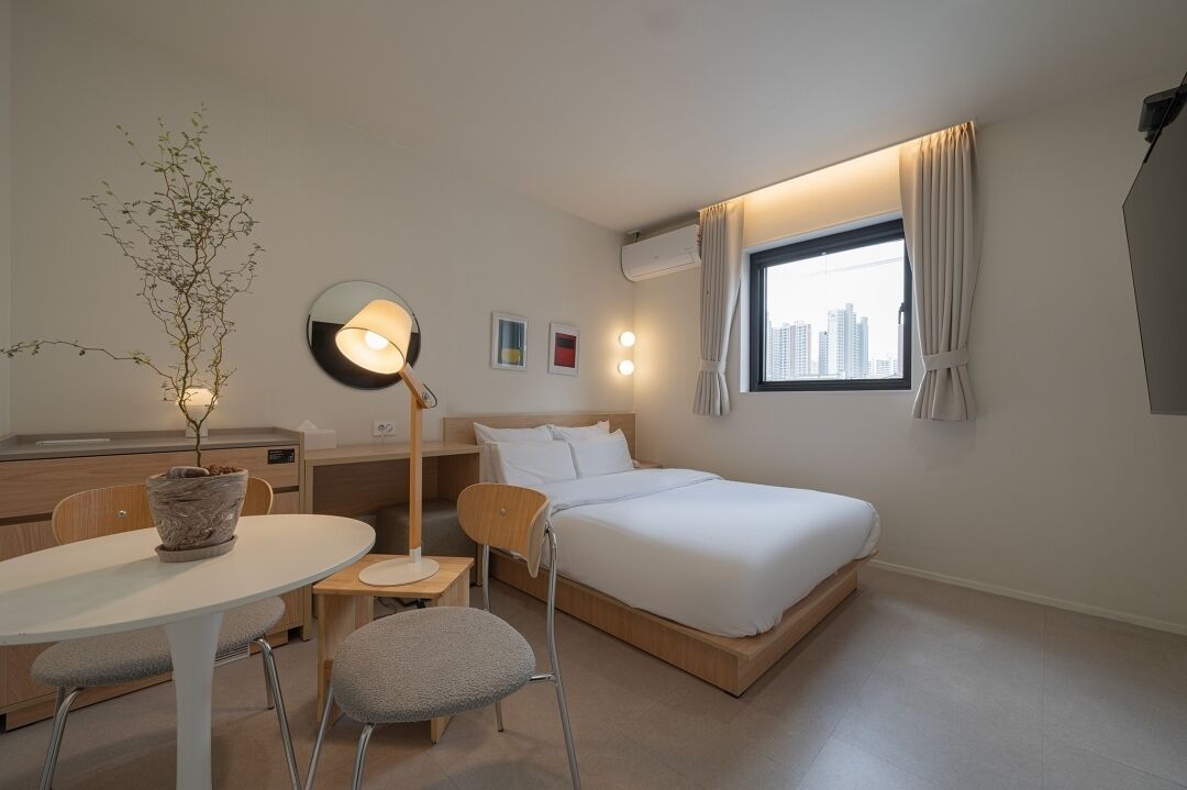 A hotel room with a bed, a desk, a chair, a table, a potted plant, a floor lamp, and a window with curtains.