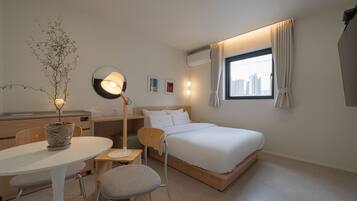 A hotel room with a bed, a desk, a chair, a table, a potted plant, a floor lamp, and a window with curtains.