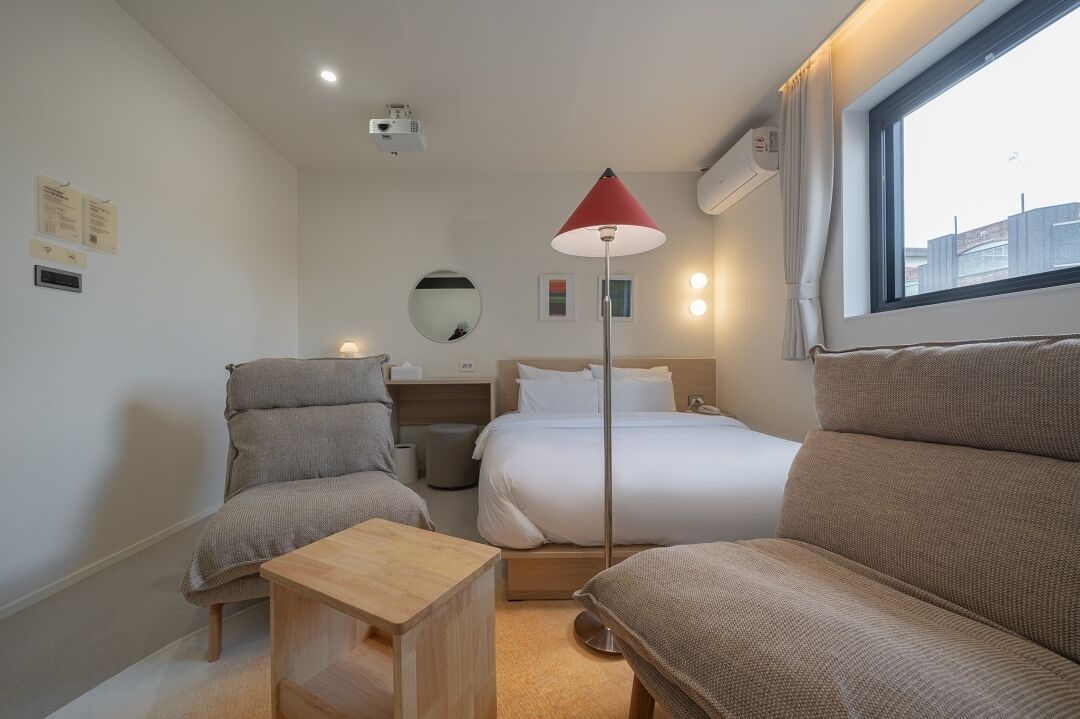 Een hotelkamer met een bed, twee fauteuils, een klein houten tafeltje, een vloerlamp, een spiegel en een raam met uitzicht op gebouwen.