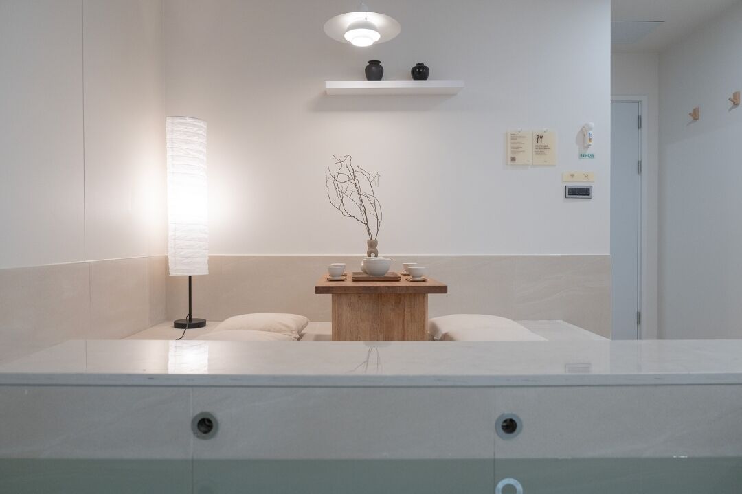 A modern bathroom with a marble vanity, a wall-mounted shelf with decorative items, a wall-mounted light fixture, and a small wooden table with a vase and cups.