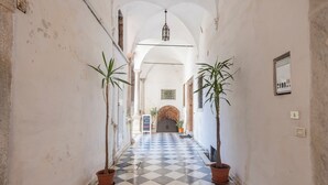 Un corridoio con pavimento a scacchi, piante in vaso e una porta centrale con un cartello.