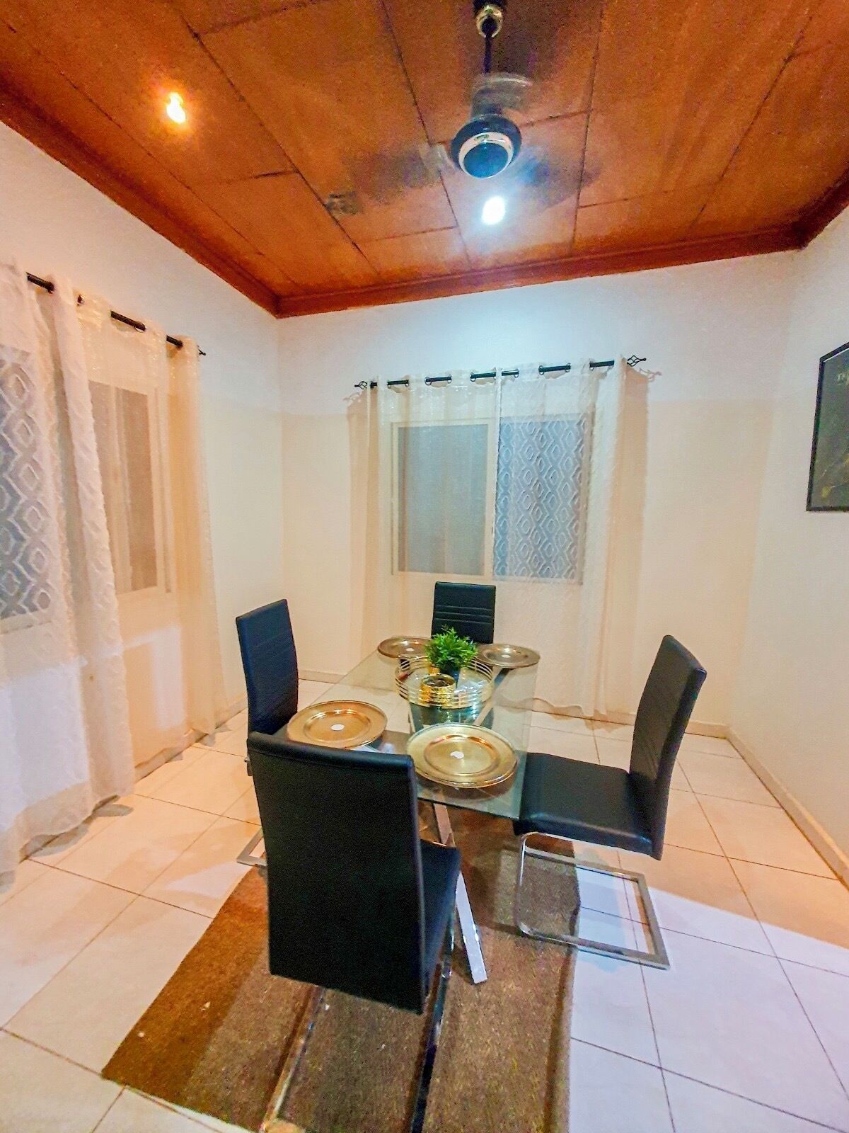 A dining area with a table set for four, surrounded by chairs, a ceiling fan, and a window with curtains.