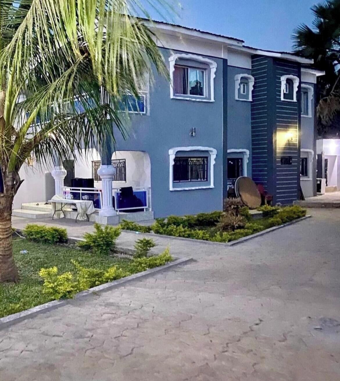 A two-story building with a flat roof, a balcony, and a small garden area with a palm tree.