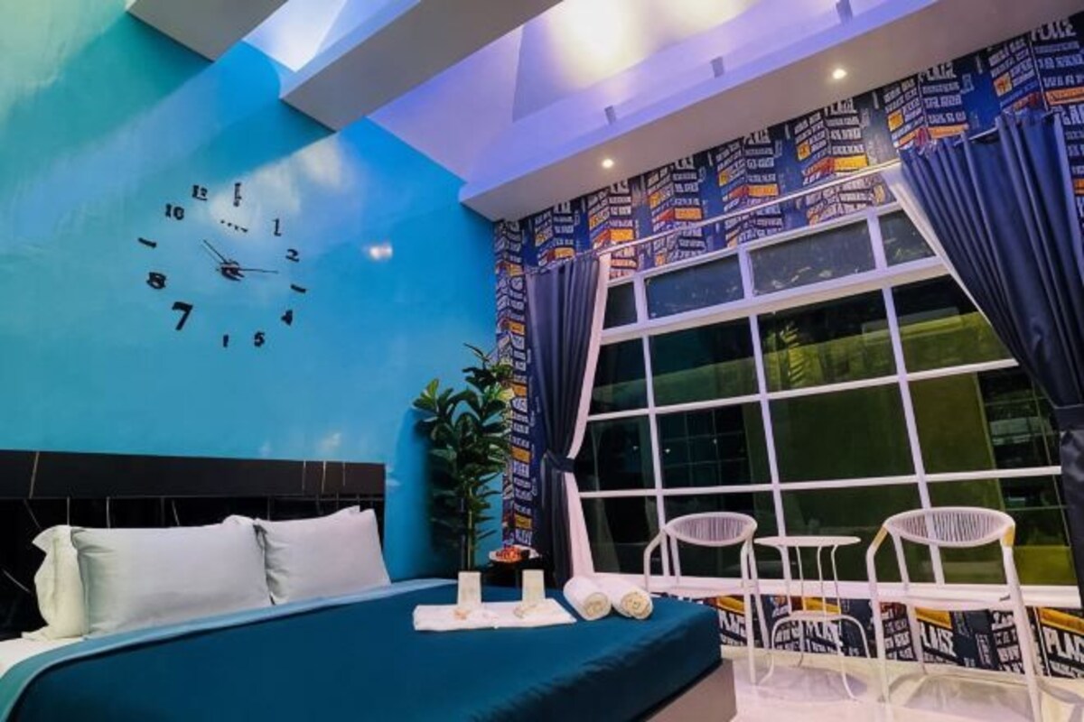 A modern hotel room with a large bed, a wall clock, a window with curtains, and a small seating area.