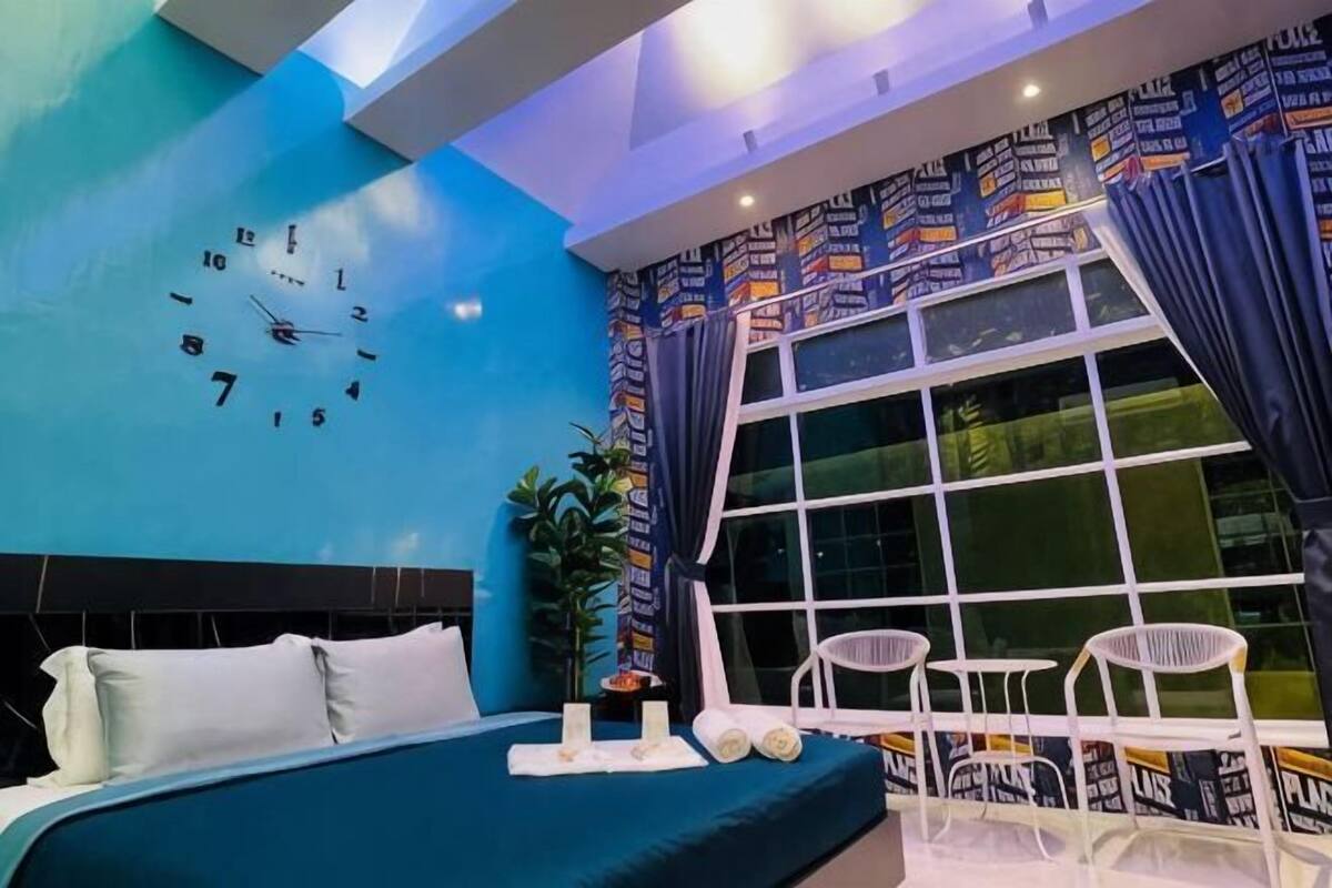 A modern hotel room with a large bed, a wall clock, a window with curtains, and a small seating area.