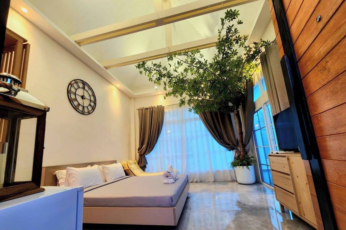A modern bedroom with a large bed, a potted plant, a wooden dresser, and a clock on the wall.