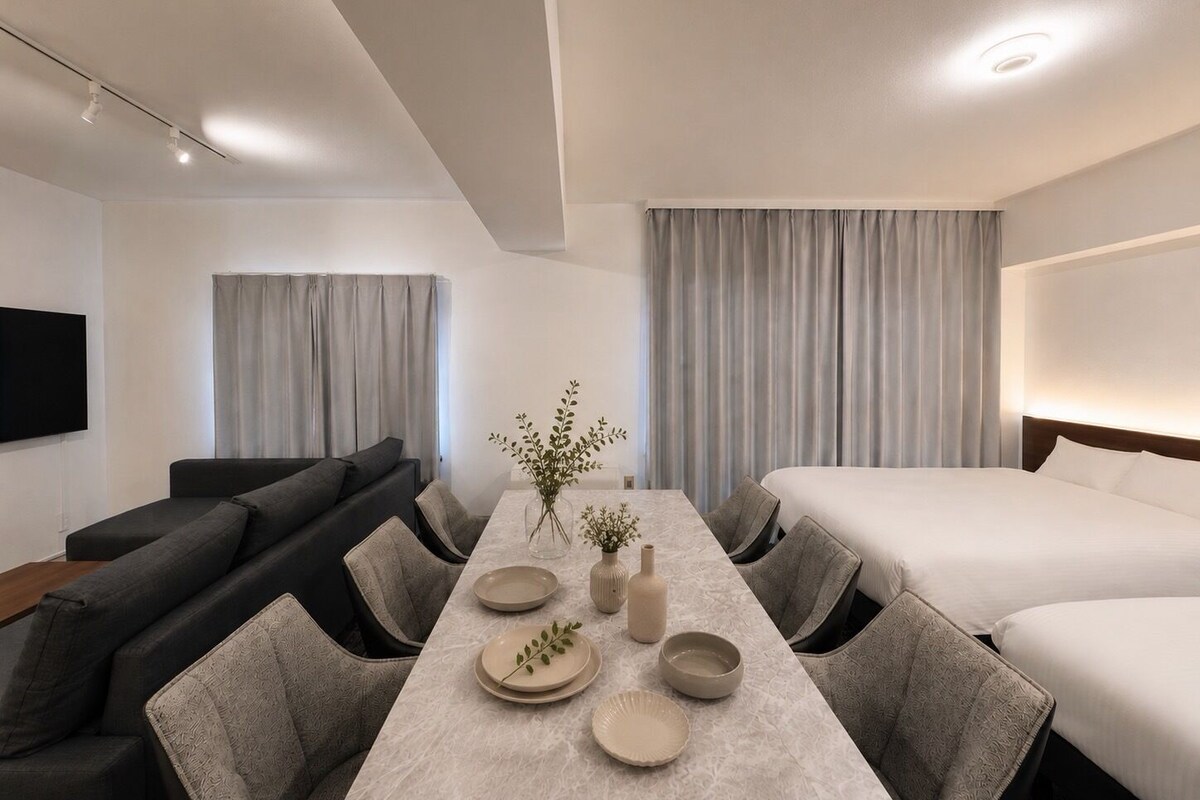 Una habitación de hotel moderna con un sofá, una cama, una mesa de comedor con sillas y un televisor montado en la pared.