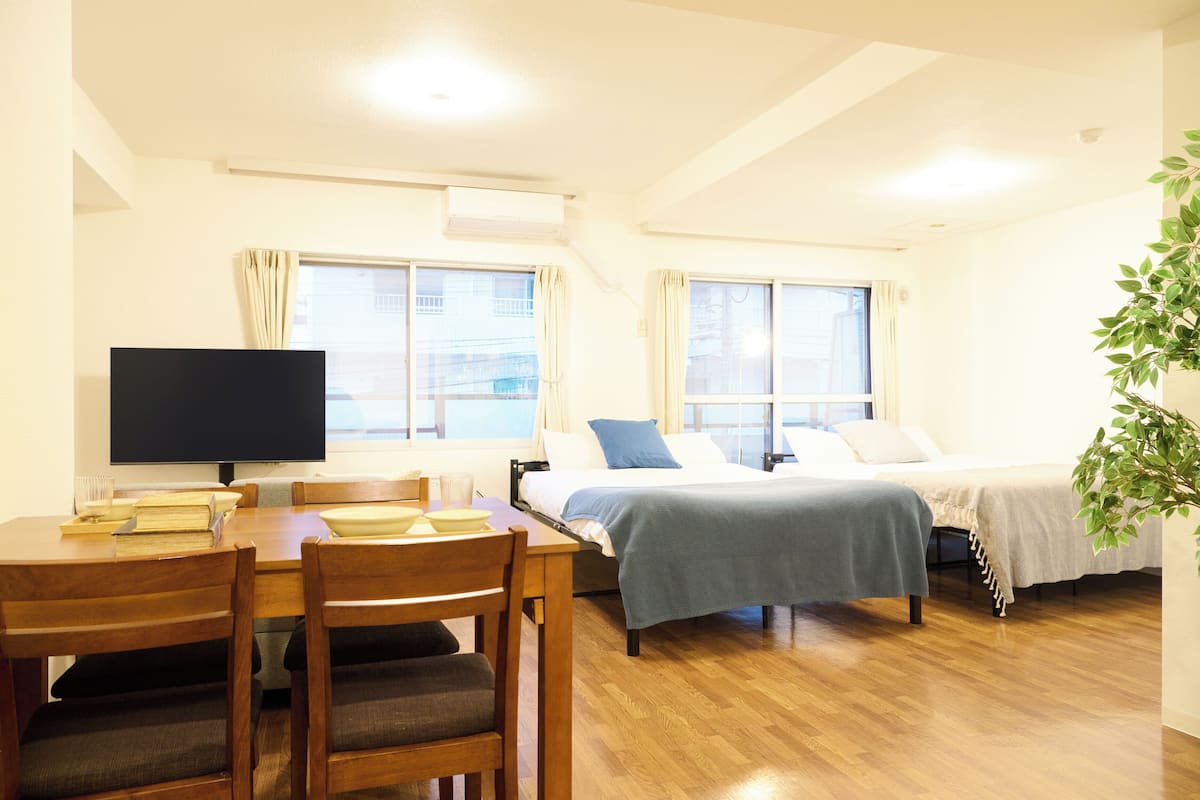 A room with a wooden dining table, chairs, a bed with a blue blanket, and a TV on a stand.