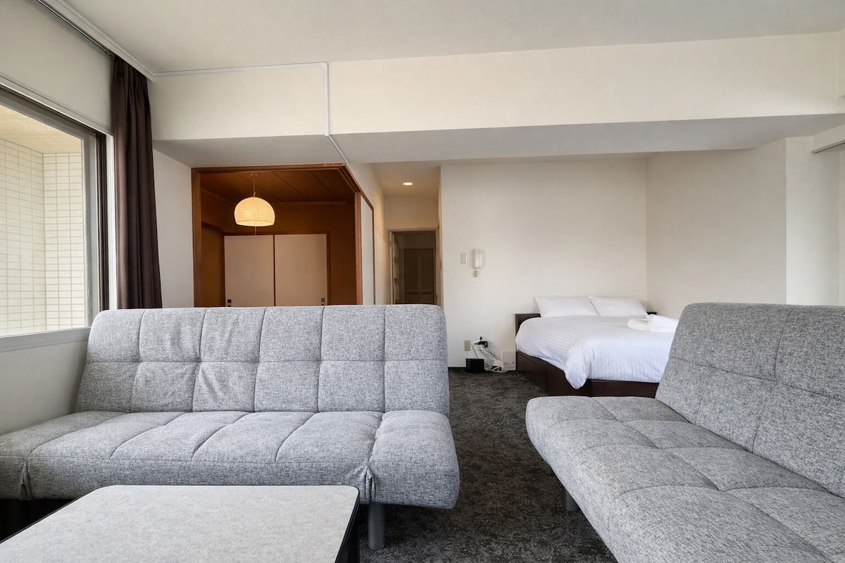A hotel room with two grey sofas, a bed, a small table, and a hanging light fixture.