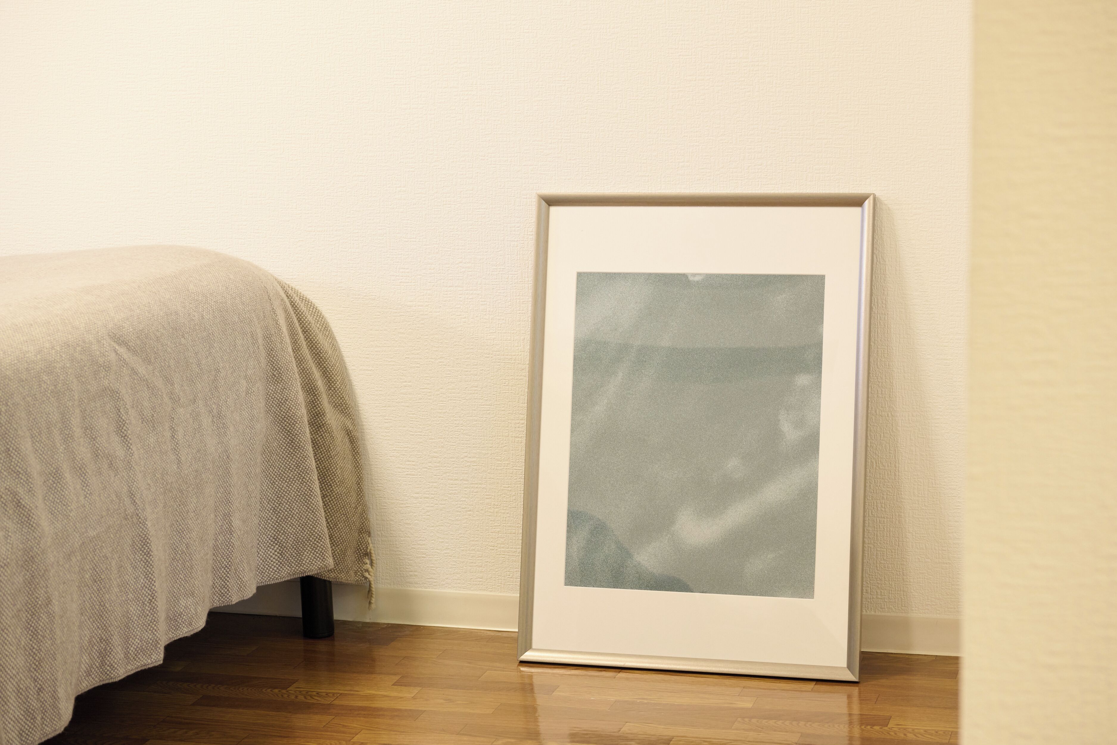 Um quadro emoldurado apoiado numa parede num quarto com soalho de madeira e cama com manta bege.