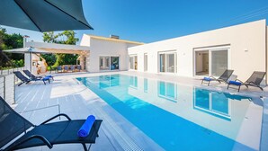 Ein moderner Außenpool mit Liegestühlen und Sonnenschirmen, umgeben von einem weißen Gebäude mit großen Fenstern und einem Ziegeldach.