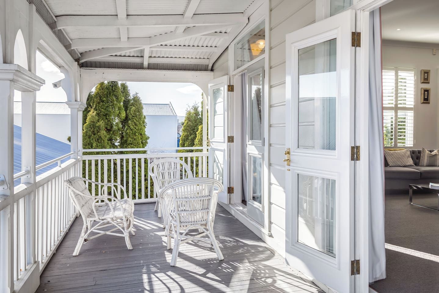 A covered porch with white railings and wicker chairs, leading to a room with a gray sofa and a coffee table.