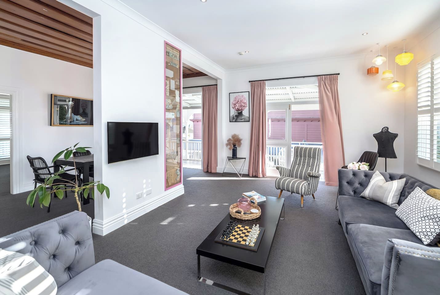 A modern living room with a grey sofa set, a coffee table with a chessboard, a TV mounted on the wall, and a large window with curtains.