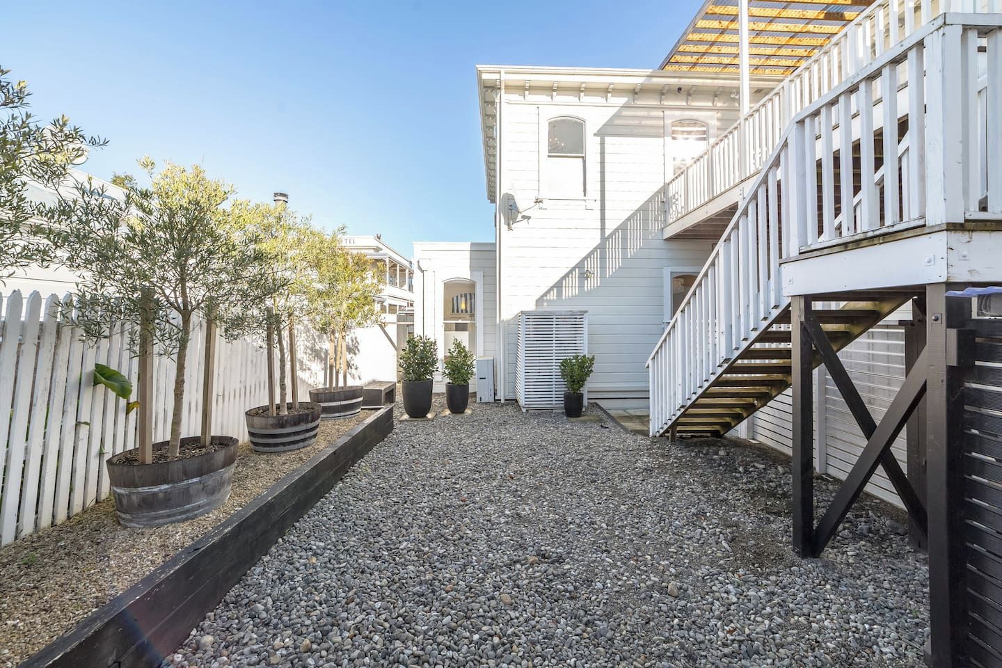 A courtyard with a gravel path, potted plants, a white fence, and a building with a staircase.