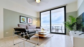 Una sala de estar moderna con un sofá, una mesa de centro y una mesa con superficie de vidrio. Hay dos cuadros en la pared, una planta y un balcón con vistas.