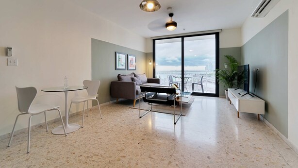 Una sala moderna con una mesa de vidrio, dos sillas blancas, un sofá y un televisor. Hay un ventanal con vistas a la ciudad.