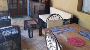 An outdoor patio area with stone flooring, wicker furniture, a wooden table with a placemat, and a lamp on a cabinet. - Belle Suite Avec Coin Cuisine Dans Maison Périgourdine (Plazac)