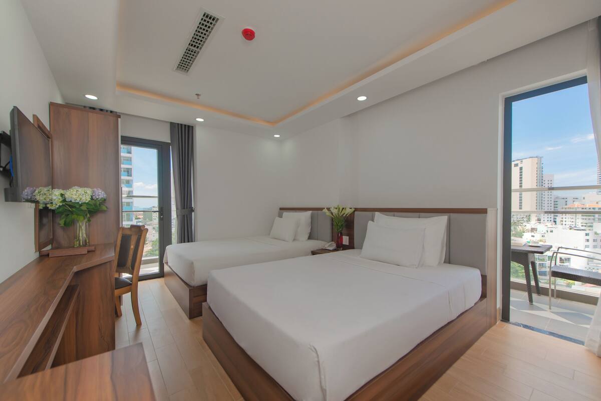 A hotel room with two beds, a desk, a chair, and a balcony with a city view.